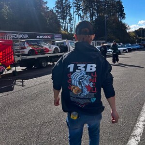 Könnte beinhalten: Eine Person trägt ein schwarzes Sweatshirt mit einem Aufdruck eines Automotors und dem Text "13B" auf dem Rücken. Die Person trägt eine blaue Jeans und eine schwarze Baseballkappe.