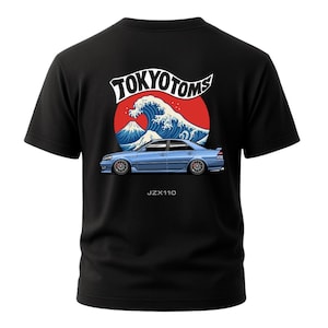 May include: Black t-shirt featuring a graphic of a blue car with a red sun, a white wave, and the text "TOKYO TOMS." Below the car is the text "JZX110." The design is on the back of the shirt.