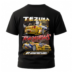 May include: Black t-shirt featuring a graphic of two yellow race cars with the text "TEZUKA" and "TOKYODRIFT" in white and red. The cars have blue and white accents and the text "GOOD YEAR". The bottom text reads "D1 GRAND PRIX SERIES".