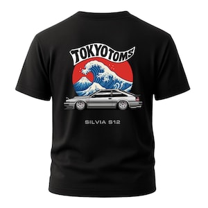 May include: Black t-shirt featuring a graphic design with the text "TOKYO TOMS" above a depiction of a wave, Mount Fuji, and a silver car. The text "SILVIA S12" is below the car.