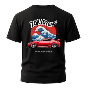 Puede incluir: Camiseta negra con un diseño de coche deportivo rojo y el texto "FAIRLADY 370Z". El diseño incluye una ola, una montaña y las palabras "TOKYO TOMS". Una camiseta gráfica para los amantes de los coches.