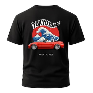 May include: Black t-shirt featuring a red convertible car graphic with the text "MIATA ND" below it. Above the car is a design with a red sun, a blue wave, and the text "TOKYO TOMS".