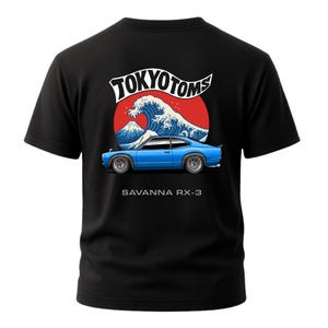 May include: Black t-shirt featuring a blue car graphic with the text "SAVANNA RX-3". The design includes a red circle, a white wave, and the text "TOKYO TOMS" in white. The shirt has short sleeves and a crew neck.