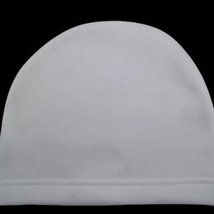 Puede incluir: Un gorro blanco liso con la parte superior redondeada y un dobladillo cosido. El gorro está hecho de un material suave y liso y está sobre un fondo negro. Este gorro sencillo y versátil es adecuado para diversas actividades.