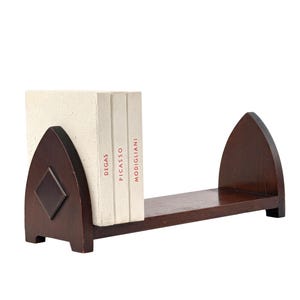 May include: A dark brown wooden bookshelf with a diamond-shaped inlay. The shelf holds three books with the names Degas, Picasso, and Modigliani printed on the spines. The bookshelf has an arched design on both ends.