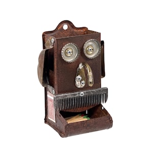 May include: A handcrafted, rusty metal match holder with a whimsical face design. The eyes are bottle caps, the nose is a curved metal piece, and the mouth is a metal comb. The matchbox drawer is open, revealing matches.