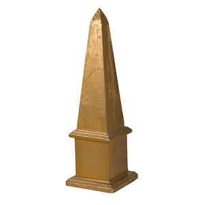 May include: A tall, gold-colored obelisk sculpture featuring a pointed pyramidal top resting on a square base. The surface has a visible, textured finish with subtle variations in the metallic gold tone across its vertical, tapered structure.