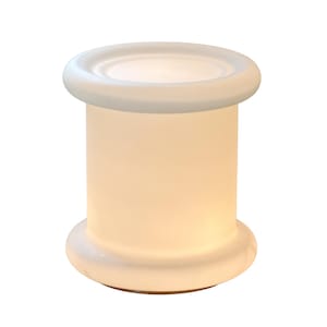 May include: A cylindrical table lamp made of frosted white glass with a smooth, matte finish. The lamp features a wide, rounded rim at both the top and the base, creating a spool-like silhouette that emits a soft, warm glow when illuminated.
