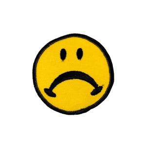 May include: A yellow, circular patch featuring a sad face design. The face has two black dots for eyes and a curved, downward-facing black mouth. The patch is outlined with a black border.