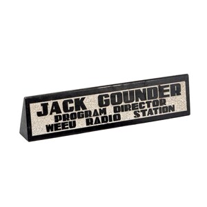 May include: A black desk nameplate with the text "JACK GOUNDER PROGRAM DIRECTOR WEEU RADIO STATION" in white lettering. The nameplate has a textured, off-white background and a black border.