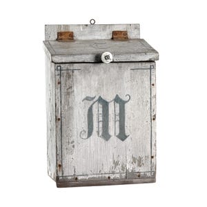 May include: A weathered gray wooden wall-mounted box with a hinged lid, a white ceramic knob, and rusty metal hinges. The front panel features a large, dark gray letter M painted in a gothic font, enclosed within a thin, dark gray rectangular border.