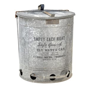 May include: A vintage Safe Guard daily waste can made of galvanized metal. The cylindrical container has a lid, handle, and air holes near the base. Text on the can reads "EMPTY EACH NIGHT" and "CELL METAL PRODUCTS CHICAGO 47 ILL."