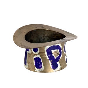 May include: A small, silver-colored metal container with a wide, oval rim and a cylindrical base. The base is decorated with blue and white painted letters. The container has a vintage or antique appearance.