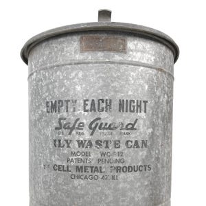 May include: A vintage galvanized steel trash can with a lid and handle. The can features the text "EMPTY EACH NIGHT" and "Safe Guard" in black lettering. Additional text includes "ILY WASTE CAN", "MODEL WC-512", and "CELL METAL PRODUCTS CHICAGO 47 ILL".