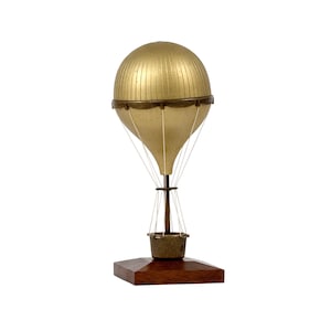May include: A gold-colored hot air balloon model with a detailed spherical top and a conical lower section. The balloon is suspended by thin white cords attached to a small basket. It is mounted on a dark brown, wooden, hexagonal base.