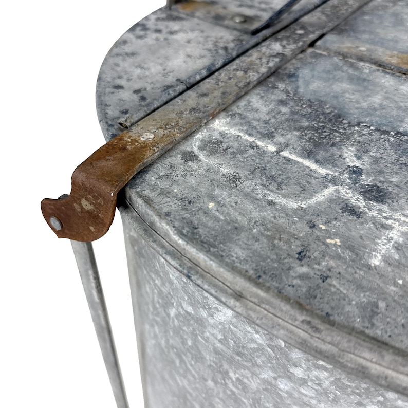 May include: Close-up of a galvanized metal container with a lid. The container has a cylindrical body and a rounded top. A rusty metal latch and handle are attached to the lid. The metal has a weathered appearance.