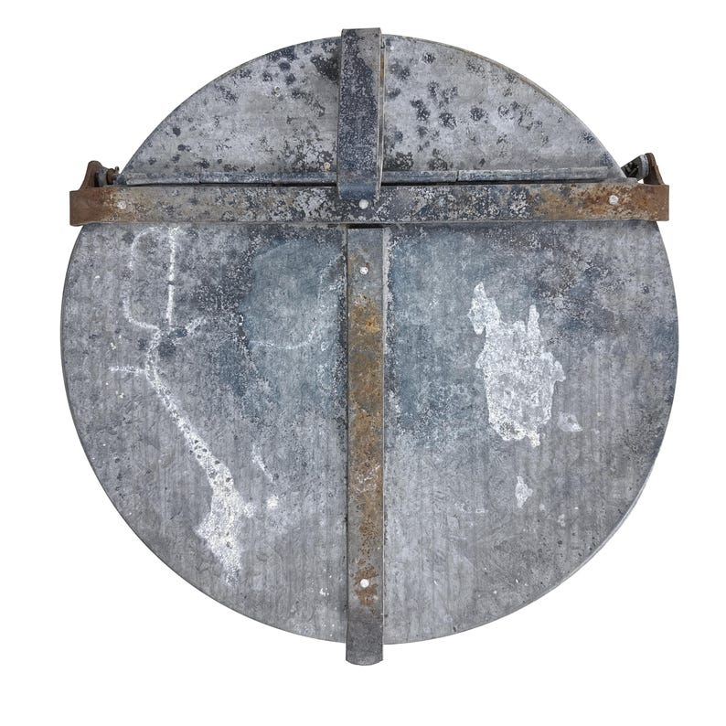 May include: A circular, weathered metal lid with a cross-shaped metal reinforcement. The lid has a distressed, mottled appearance with rust-colored accents on the metal straps. The design suggests a vintage or industrial aesthetic.