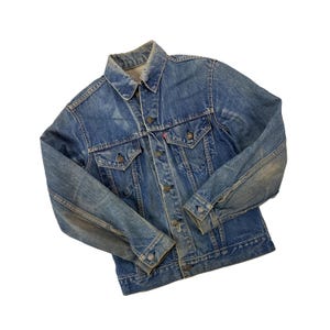 May include: A vintage blue denim jacket with a button-up front, two chest pockets, and buttoned cuffs. The jacket has a classic collar and a red tag on the chest pocket. The denim shows signs of wear, giving it a distressed look.