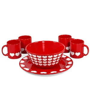 May include: A red ceramic dinnerware set featuring a large bowl, a plate, and four mugs. The set is decorated with white heart patterns. The mugs have handles and two white hearts. The bowl and plate have a border of white hearts.