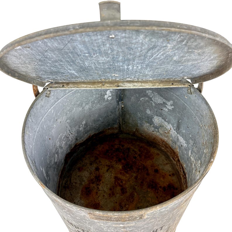 May include: An open, galvanized metal bucket with a hinged lid. The bucket has a cylindrical shape with a slightly tapered body. The interior shows signs of rust and wear, suggesting age and use. A small handle is visible on the lid.