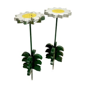 May include: Two wooden daisy flower garden stakes. Each stake has a white and yellow flower head, a green stem, and green leaves. The paint has a distressed, vintage look.