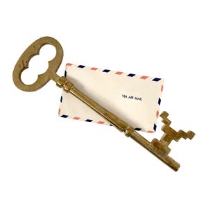 May include: A large, antique-style brass key with a decorative handle rests atop a white envelope. The envelope is trimmed with pink and blue stripes and features the text "VIA AIR MAIL".