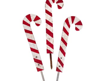 May include: Three wooden candy cane decorations featuring a distressed white and red striped pattern. Each piece has a curved top and a pointed metal stake at the bottom for insertion into the ground, showcasing a weathered, rustic finish.