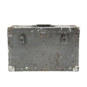 May include: A weathered, gray metal trunk with a handle and two latches. The trunk shows signs of age with paint chipping and rust-like stains. The corners are reinforced with metal plates.