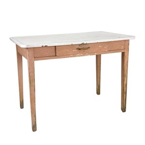 May include: A vintage desk with a white tabletop and a single drawer. The desk has a distressed, pinkish-brown painted frame and legs. The drawer has a wooden handle. The desk is approximately 40 inches long.