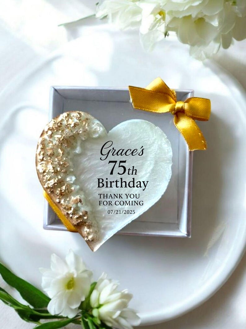 May include: A heart-shaped gift in a white box, with gold accents on one side. The heart has the text "Grace's 75th Birthday" and "Thank you for coming 07/21/2025". A gold ribbon is tied on the box.