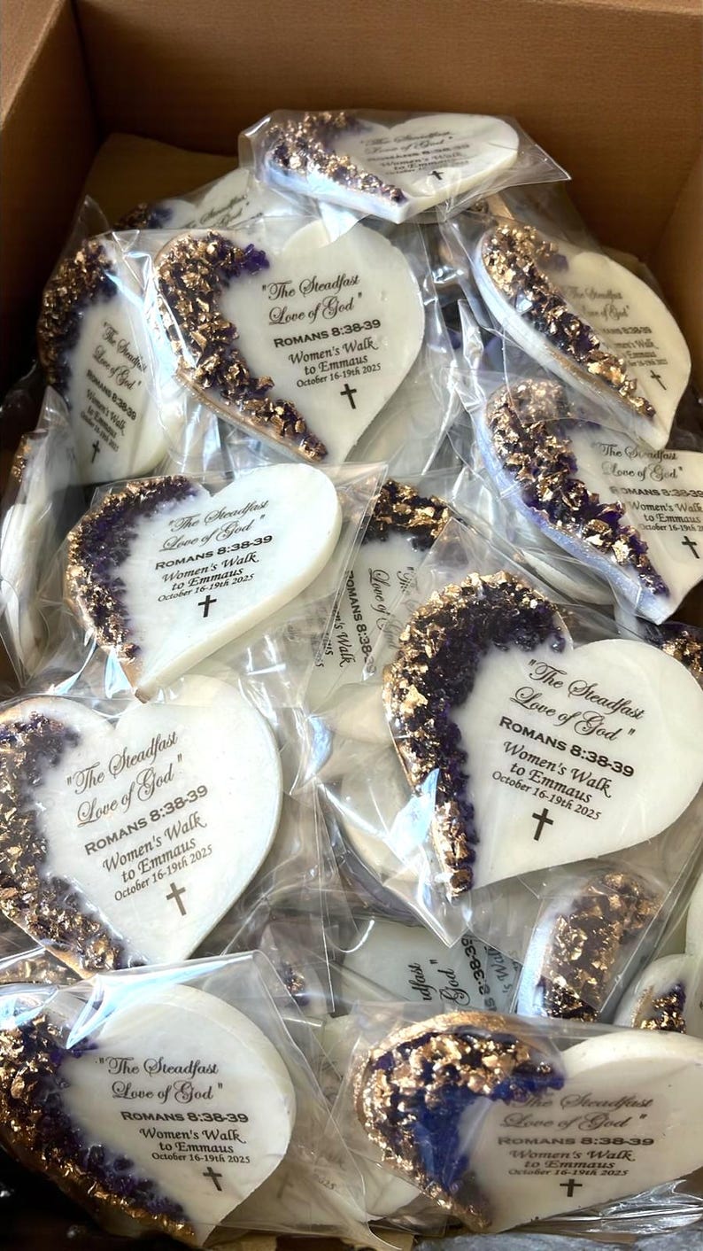 May include: Heart-shaped cookies with a white base and a decorative edge of gold and purple accents. Each cookie is individually wrapped and features the text "The Steadfast Love of God" and other text.