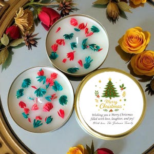 May include: Three round candles with white wax and colourful accents. Two candles have red and teal accents. The third candle has a lid with a Christmas tree design and the text "Merry Christmas". The candles are on a reflective surface with floral decorations.