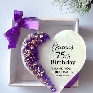 May include: A heart-shaped gift in a box. The left side is purple with gold flakes, the right side is white. Text reads "Grace's 75th Birthday, Thank you for coming 07/21/2025". A purple ribbon is tied on the box.