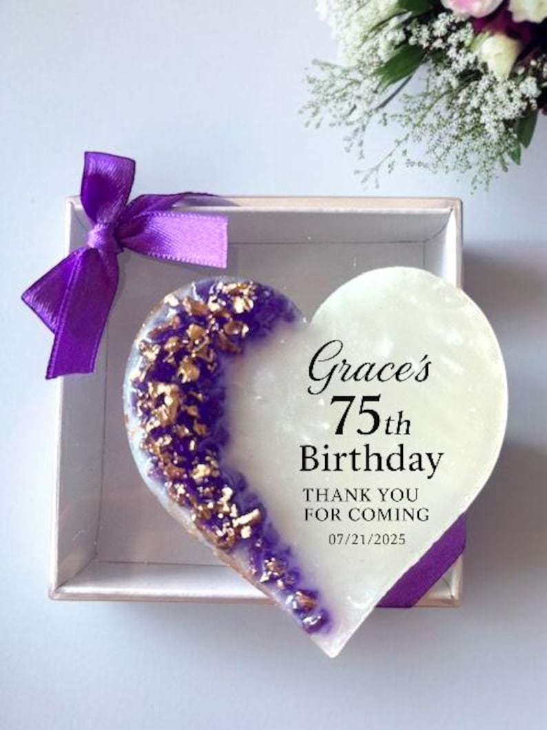 May include: A heart-shaped gift in a box. The left side is purple with gold flakes, the right side is white. Text reads "Grace's 75th Birthday, Thank you for coming 07/21/2025". A purple ribbon is tied on the box.