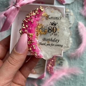 Pode incluir: Uma lembrancinha de resina para um 80&ordm; anivers&aacute;rio. A lembrancinha &eacute; branca com uma borda rosa e dourada, e o texto diz "80&ordm; Anivers&aacute;rio de Loreena Obrigado por vir". A lembrancinha est&aacute; em uma caixa branca com uma fita rosa.