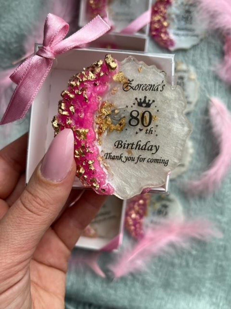 Pode incluir: Uma lembrancinha de resina para um 80&ordm; anivers&aacute;rio. A lembrancinha &eacute; branca com uma borda rosa e dourada, e o texto diz "80&ordm; Anivers&aacute;rio de Loreena Obrigado por vir". A lembrancinha est&aacute; em uma caixa branca com uma fita rosa.