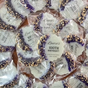 Pode incluir: Uma cole&ccedil;&atilde;o de biscoitos embalados individualmente. Cada biscoito &eacute; branco com uma borda decorativa roxa e dourada. O texto "Laura's 100th Birthday" est&aacute; impresso em cada biscoito. Os biscoitos s&atilde;o embalados em pl&aacute;stico transparente.
