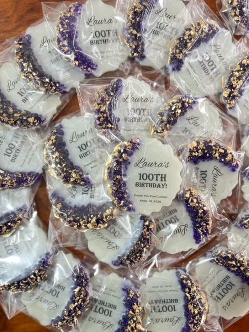 Pode incluir: Uma cole&ccedil;&atilde;o de biscoitos embalados individualmente. Cada biscoito &eacute; branco com uma borda decorativa roxa e dourada. O texto "Laura's 100th Birthday" est&aacute; impresso em cada biscoito. Os biscoitos s&atilde;o embalados em pl&aacute;stico transparente.