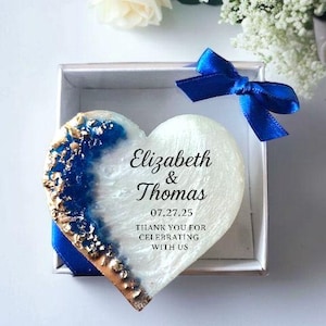 May include: A heart-shaped keepsake with blue and gold accents, presented in a silver box with a blue ribbon. The keepsake has the names "Elizabeth & Thomas" and the date "07.27.25" with the text "THANK YOU FOR CELEBRATING WITH US". White flowers are in the background.