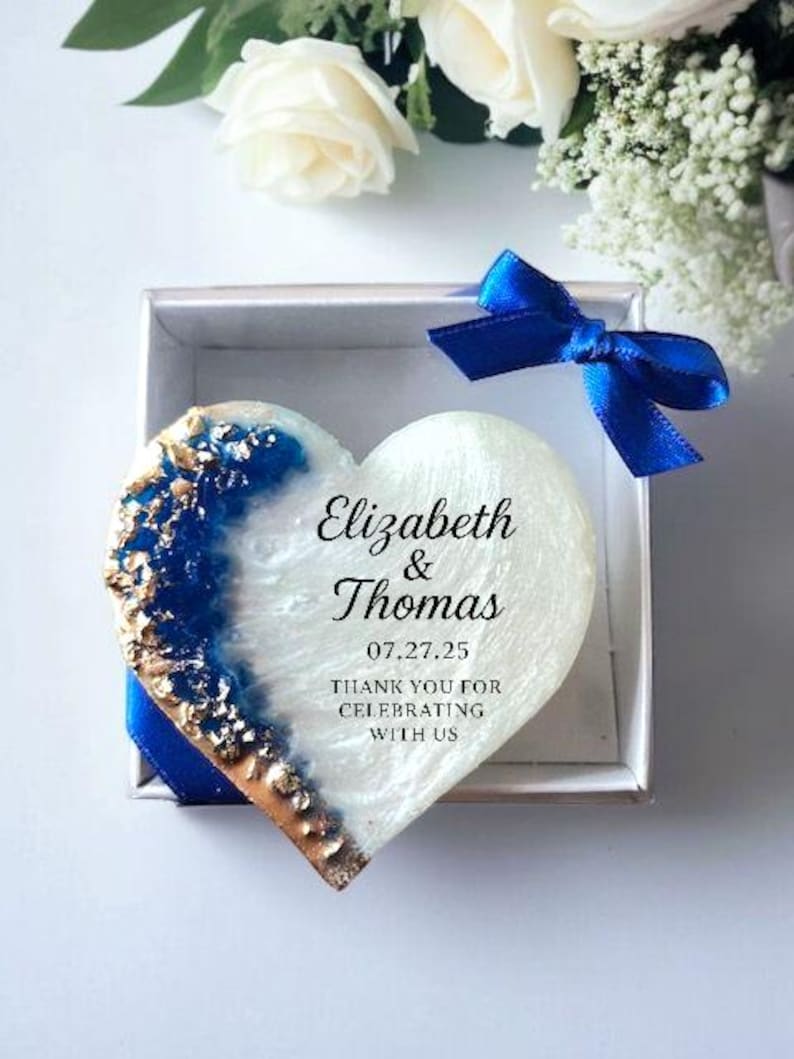 May include: A heart-shaped keepsake with blue and gold accents, presented in a silver box with a blue ribbon. The keepsake has the names "Elizabeth & Thomas" and the date "07.27.25" with the text "THANK YOU FOR CELEBRATING WITH US". White flowers are in the background.