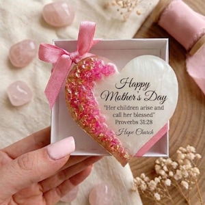 May include: A heart-shaped Mother's Day gift in a white box, adorned with a pink ribbon. The heart is half-filled with pink and gold glitter, and the other half has the text "Happy Mother's Day" and a Bible verse. Pink gemstones are scattered around.