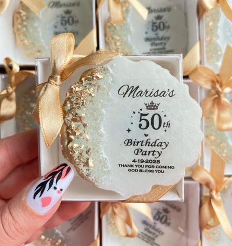 Pode incluir: Uma lembrancinha de festa branca, de formato irregular, com detalhes dourados e o texto "Marisa's 50th Birthday Party 4-19-2025". A lembrancinha est&aacute; em uma caixa branca com uma fita dourada.