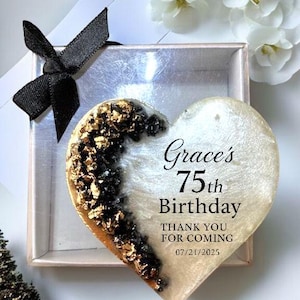 May include: A heart-shaped keepsake with gold and black accents, inscribed with "Grace's 75th Birthday." The keepsake is presented in a small box with a black bow. White flowers are in the background.