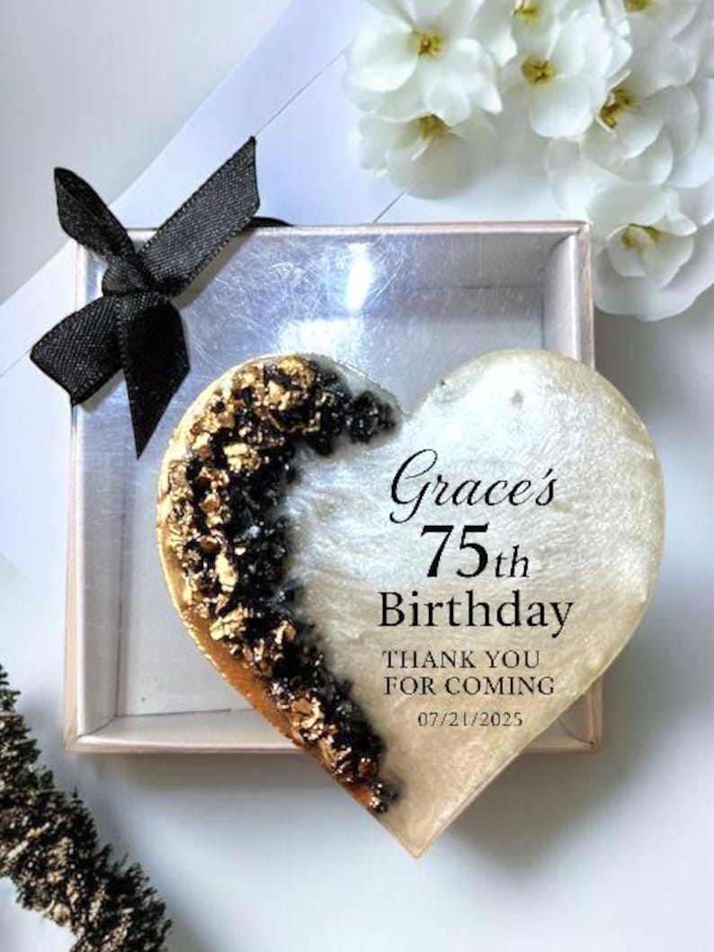 May include: A heart-shaped keepsake with gold and black accents, inscribed with "Grace's 75th Birthday." The keepsake is presented in a small box with a black bow. White flowers are in the background.