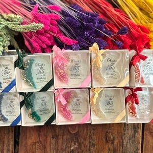 Pode incluir: Caixas de presente com itens decorativos com glitter, amarradas com fitas coloridas. O texto "Frisks 55th Birthday" est&aacute; impresso em cada caixa. Flores secas coloridas no fundo.