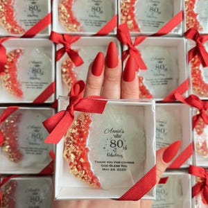 Pode incluir: Uma cole&ccedil;&atilde;o de caixas quadradas brancas, cada uma contendo uma base decorativa com detalhes vermelhos e dourados. As bases est&atilde;o inscritas com "Annie's 80th & Fabulous" e detalhes do evento. Cada caixa &eacute; amarrada com uma fita vermelha.