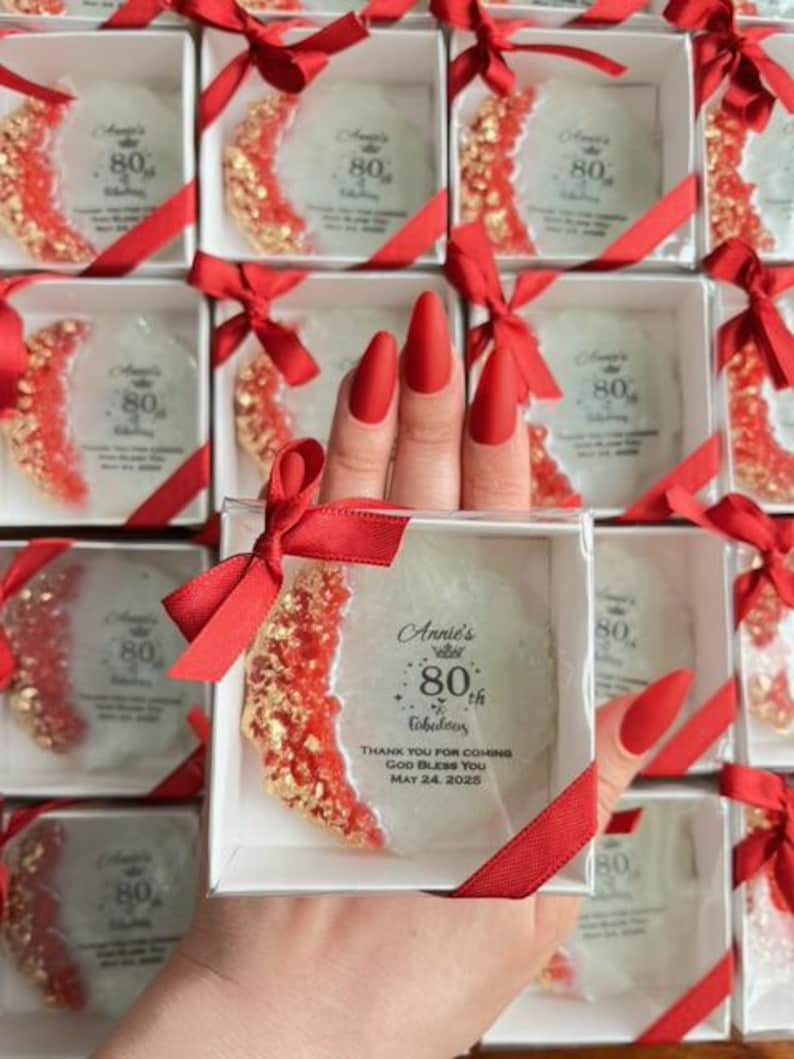 Pode incluir: Uma cole&ccedil;&atilde;o de caixas quadradas brancas, cada uma contendo uma base decorativa com detalhes vermelhos e dourados. As bases est&atilde;o inscritas com "Annie's 80th & Fabulous" e detalhes do evento. Cada caixa &eacute; amarrada com uma fita vermelha.