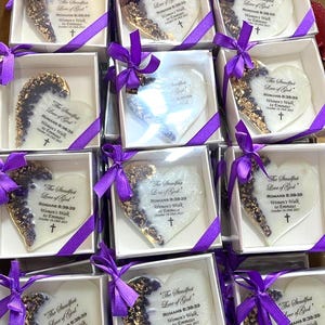 May include: Heart-shaped keepsakes in white boxes, adorned with purple ribbons. Each heart has a gold-leaf design on one side and text that reads "The Steadfast Love of God" and other text. A cross is also visible.