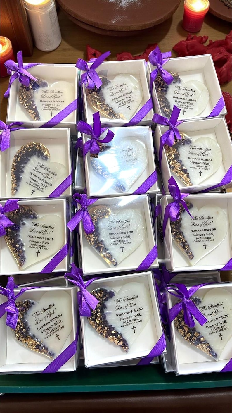 May include: Heart-shaped keepsakes in white boxes, adorned with purple ribbons. Each heart has a gold-leaf design on one side and text that reads "The Steadfast Love of God" and other text. A cross is also visible.