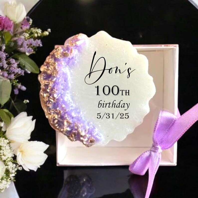 Pode incluir: Um objeto decorativo em forma de geodo branco sujo com uma borda roxa e dourada, apresentado numa caixa branca com uma fita roxa. O texto "Don's 100th birthday 5/31/25" est&aacute; escrito no objeto. Flores no fundo.